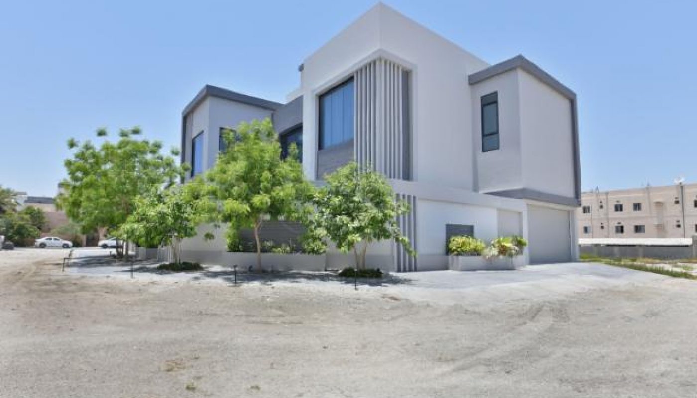 Modern white two-story house with large windows, surrounded by a few small green trees, situated on a bare gravel lot under a clear blue sky.