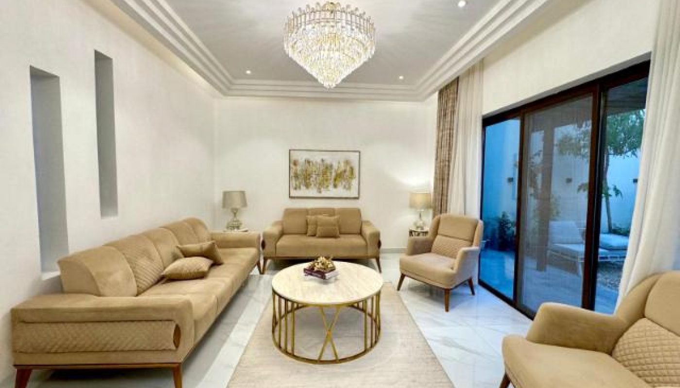Modern living room with beige sofas and armchairs, round coffee table, chandelier, neutral decor, and large glass doors leading outside.