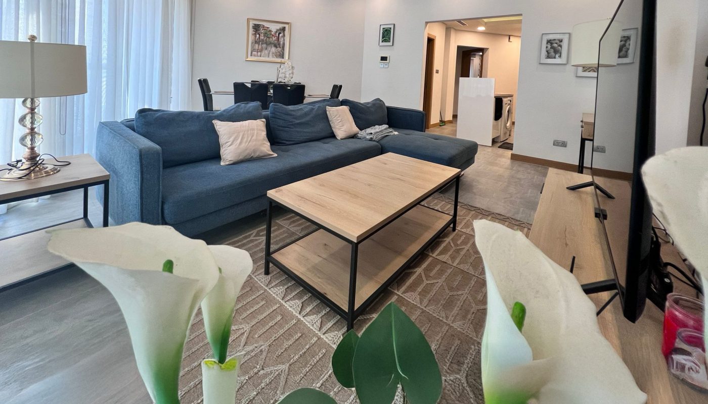 Modern living room with a blue sofa, wooden coffee table, and decorative white calla lilies in the foreground. Dining area and artwork visible in the background. Natural light through sheer curtains.