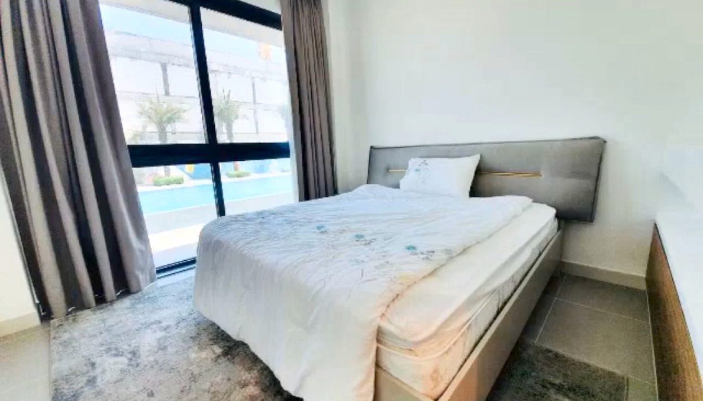 Bright bedroom with a double bed, gray headboard, large window with curtains, and a view of a pool and palm trees outside.