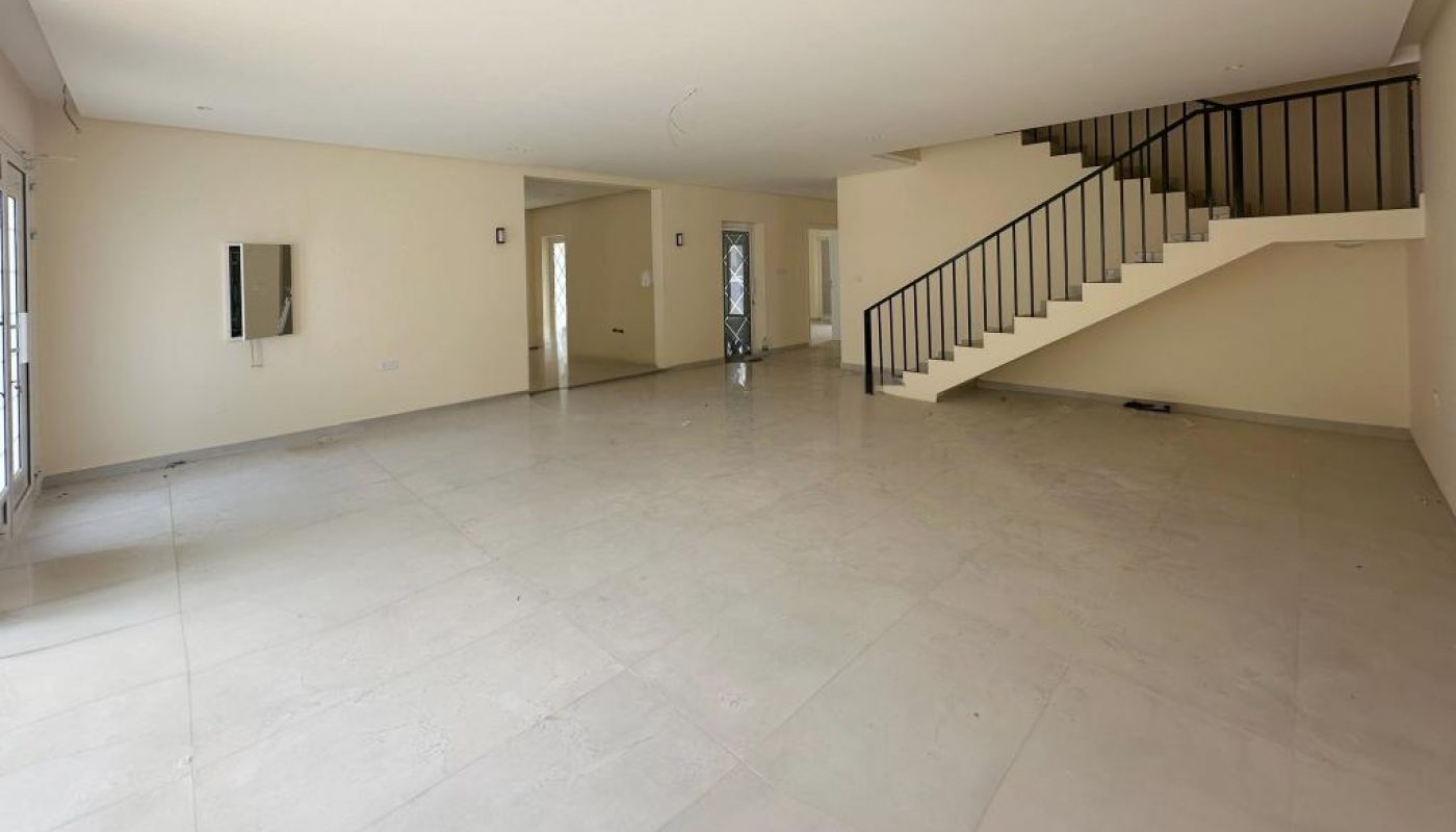 Spacious empty room with tiled floors, beige walls, and a staircase with a black railing leading to an upper floor. Natural light enters through windows on the left.