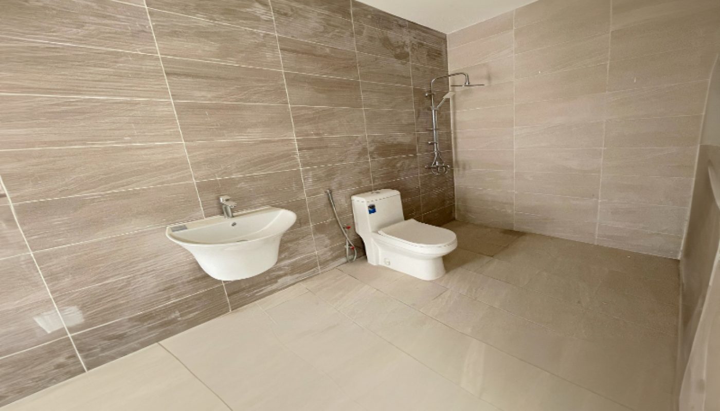 Spacious bathroom with light brown wall tiles, white floor tiles, a wall-mounted sink, a toilet, and an open shower area.