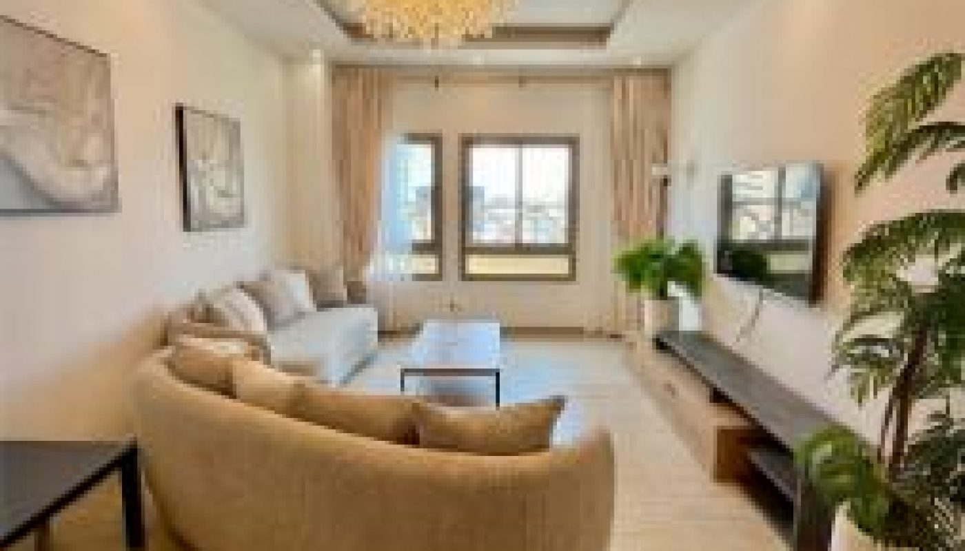 Modern living room with a beige curved sofa, wall art, chandelier, large window, potted plants, and a wooden TV console on tiled flooring.