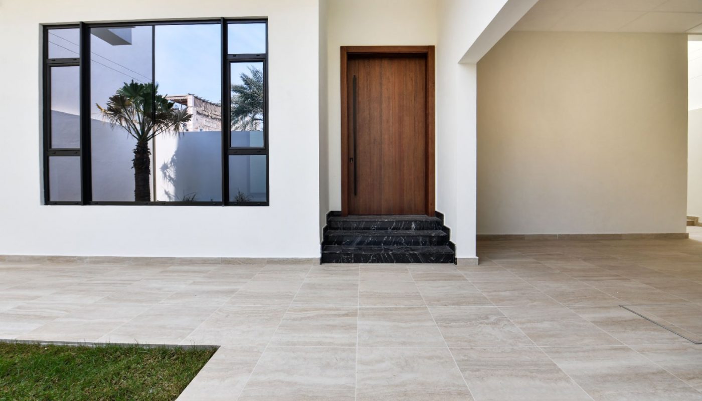 Modern entryway with a wooden door, large window, and tiled floor. Small patch of grass to the left. White walls and ceiling create a clean, minimalist look.