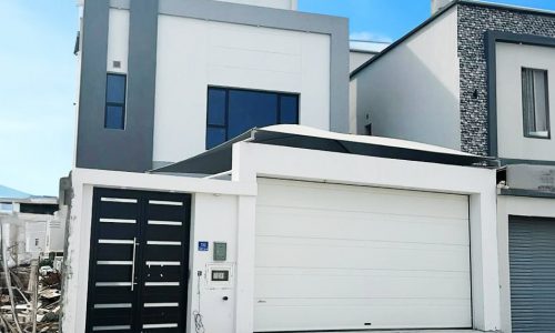 Modern 3BR villa with a gray and white facade, featuring a large garage door and a contemporary front door, under a clear blue sky.