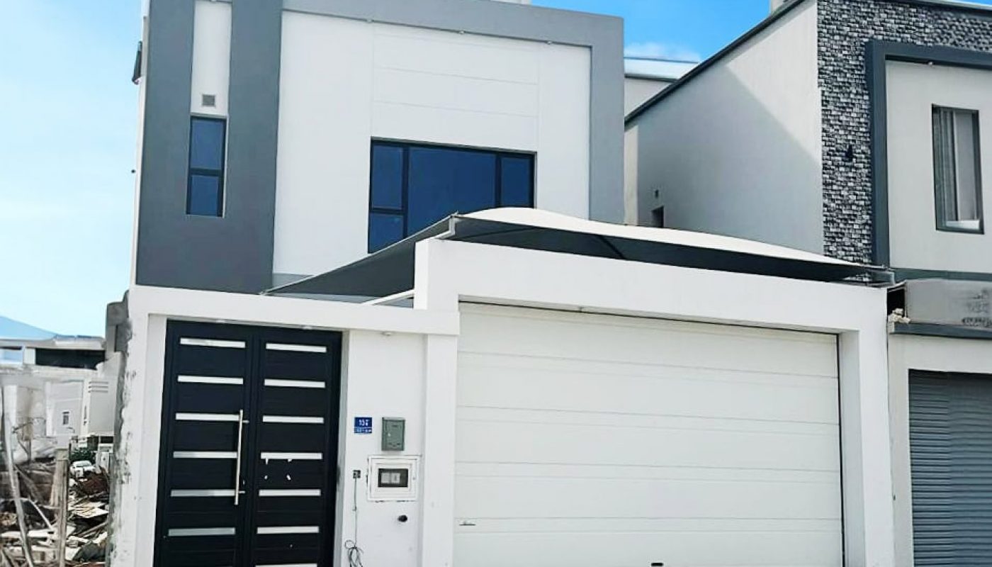 Modern 3BR villa with a gray and white facade, featuring a large garage door and a contemporary front door, under a clear blue sky.