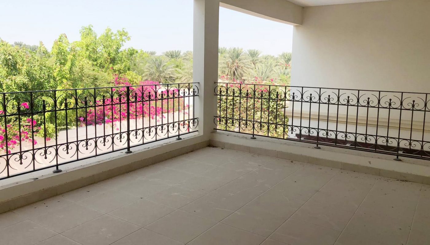 An empty covered balcony with tiled flooring and iron railings, offering a serene view of trees and blooming flowers in the distance – just one of the many charming features of this elegant 4BR SF Villa in Hamala.