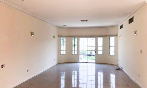 A large empty room with white walls, tiled floor, ceiling lights, and a set of French doors leading to a garden.