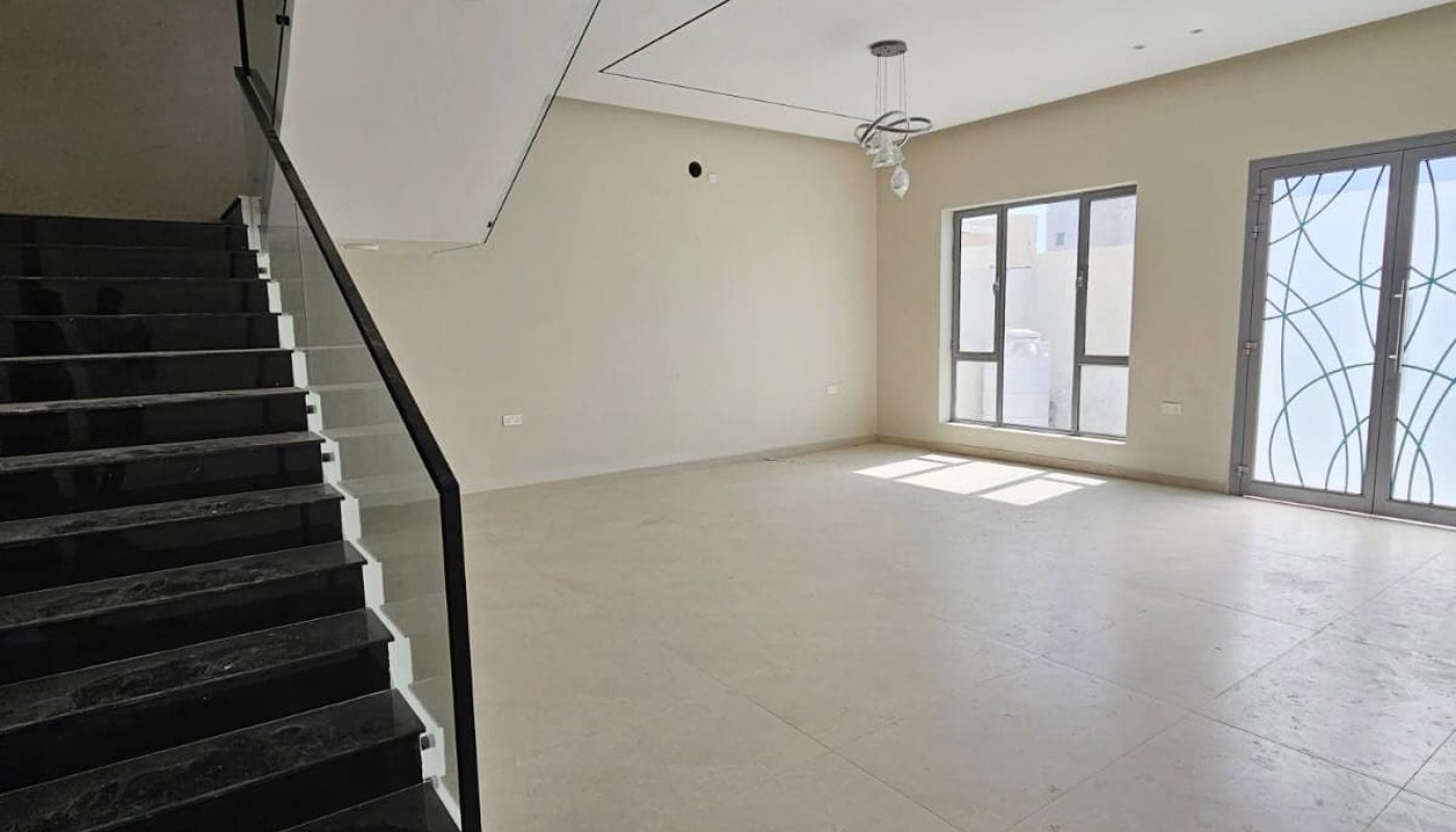 A modern, unfurnished room with a staircase on the left, large windows with decorative patterns on the right, and a light fixture hanging from the ceiling.