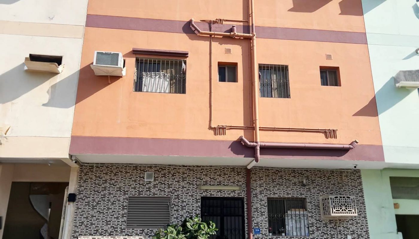 A small, three-story building with an orange upper facade and tiled lower section. Air conditioning units are visible under the windows. A plant grows beside the entrance, and a clear, blue sky is above.