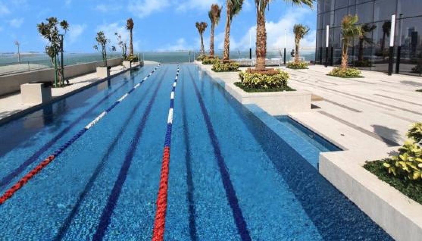 A modern outdoor swimming pool with lane dividers, surrounded by palm trees and a building, under a partly cloudy sky.