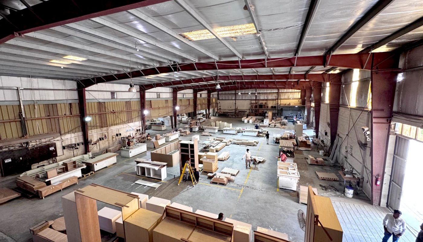 A wide view of an industrial warehouse interior shows workers assembling and organizing wooden furniture and cabinetry under bright overhead lighting.