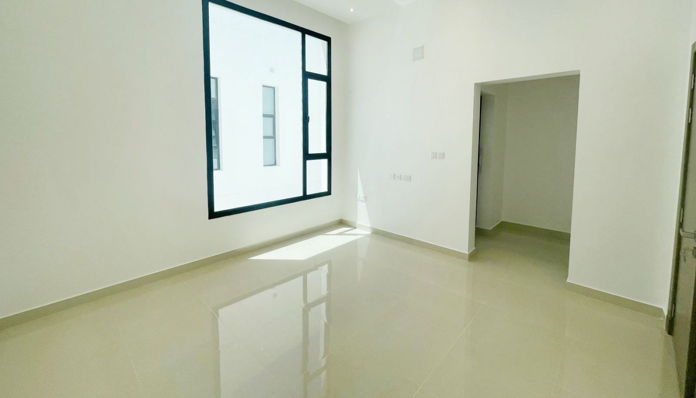 Empty, unfurnished room with beige tiled floors, white walls, a large window letting in natural light, and an open doorway leading to another area.