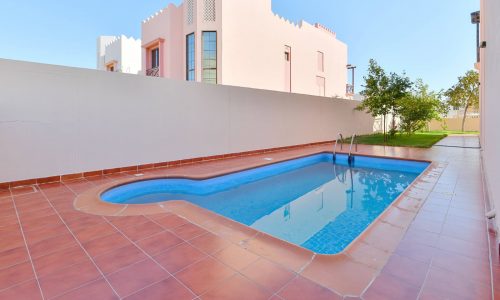 A small backyard swimming pool with a ladder, surrounded by a red-tiled deck, next to a white wall and adjacent to a beautiful 3BR Villa in the Al Areen community.