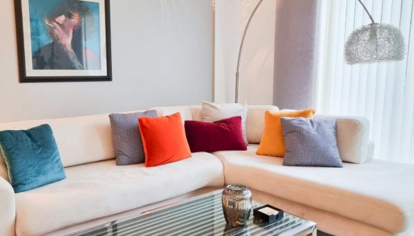 A modern living room with a white sectional sofa, colorful throw pillows, a glass coffee table, a red patterned rug, wall art, and a floor lamp near a window with vertical blinds.
