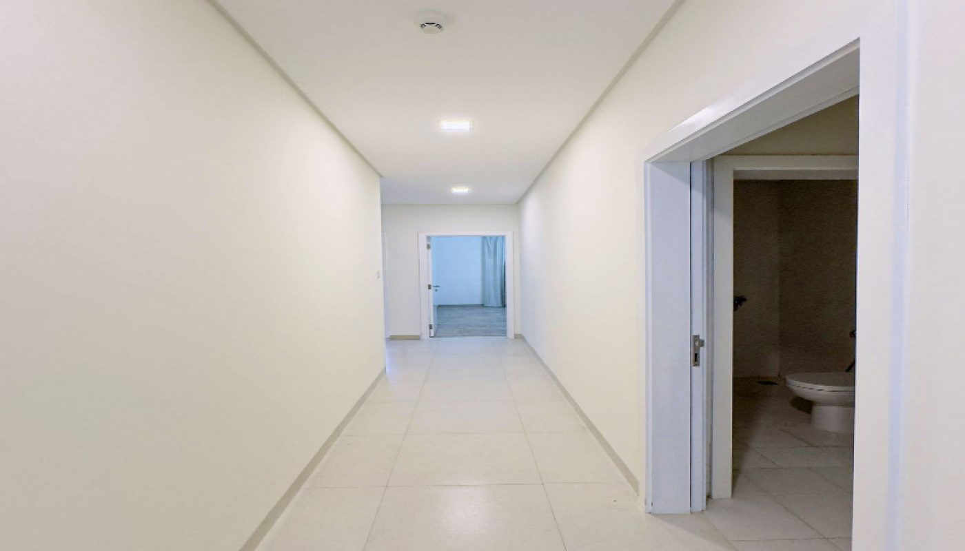 A white hallway with tiled floors, bright overhead lights, and open doors on the right side; a room with blue curtains is visible at the far end.