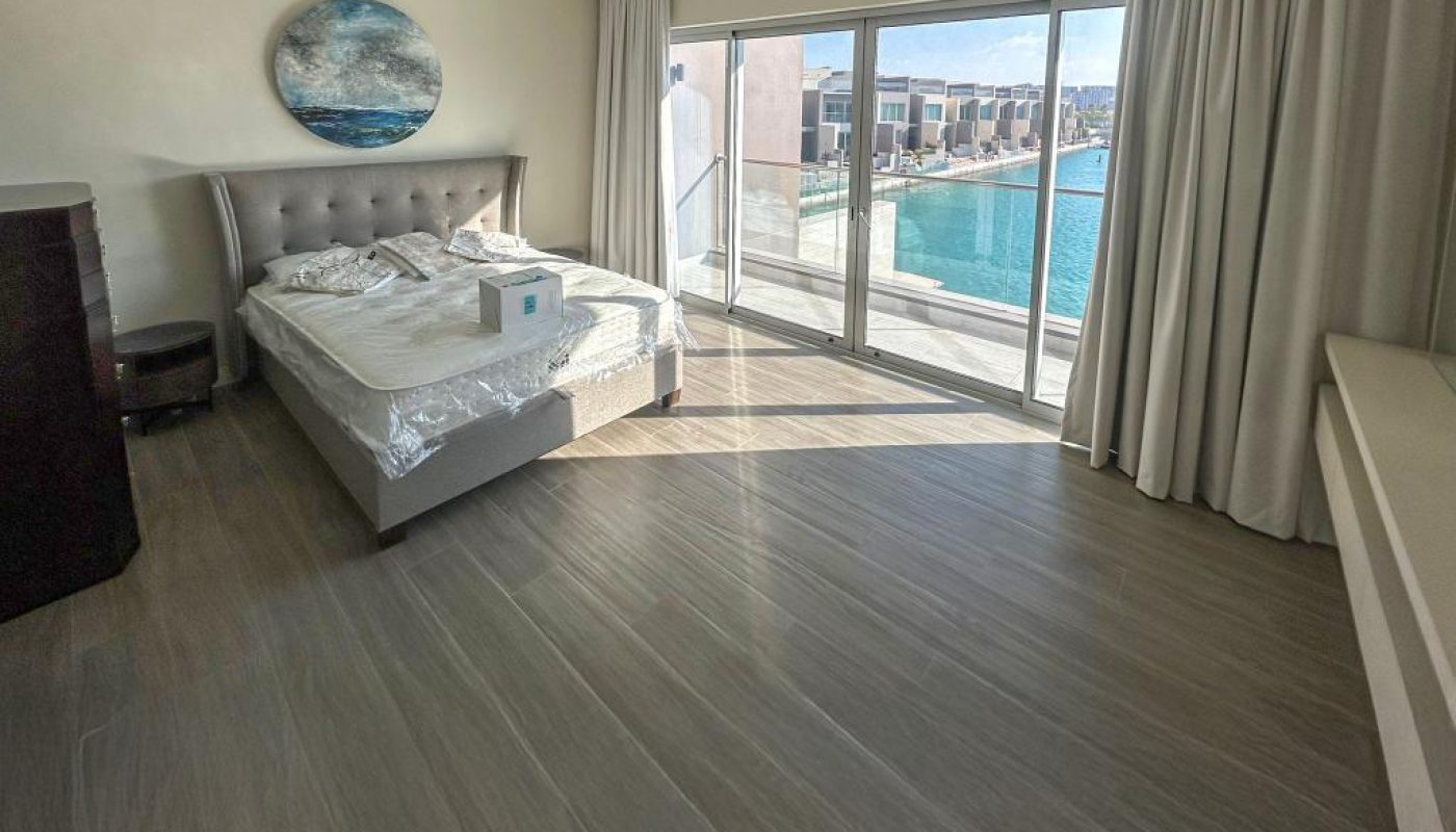 A modern bedroom with a queen bed, large windows, beige curtains, light wood flooring, and a view of a pool outside.