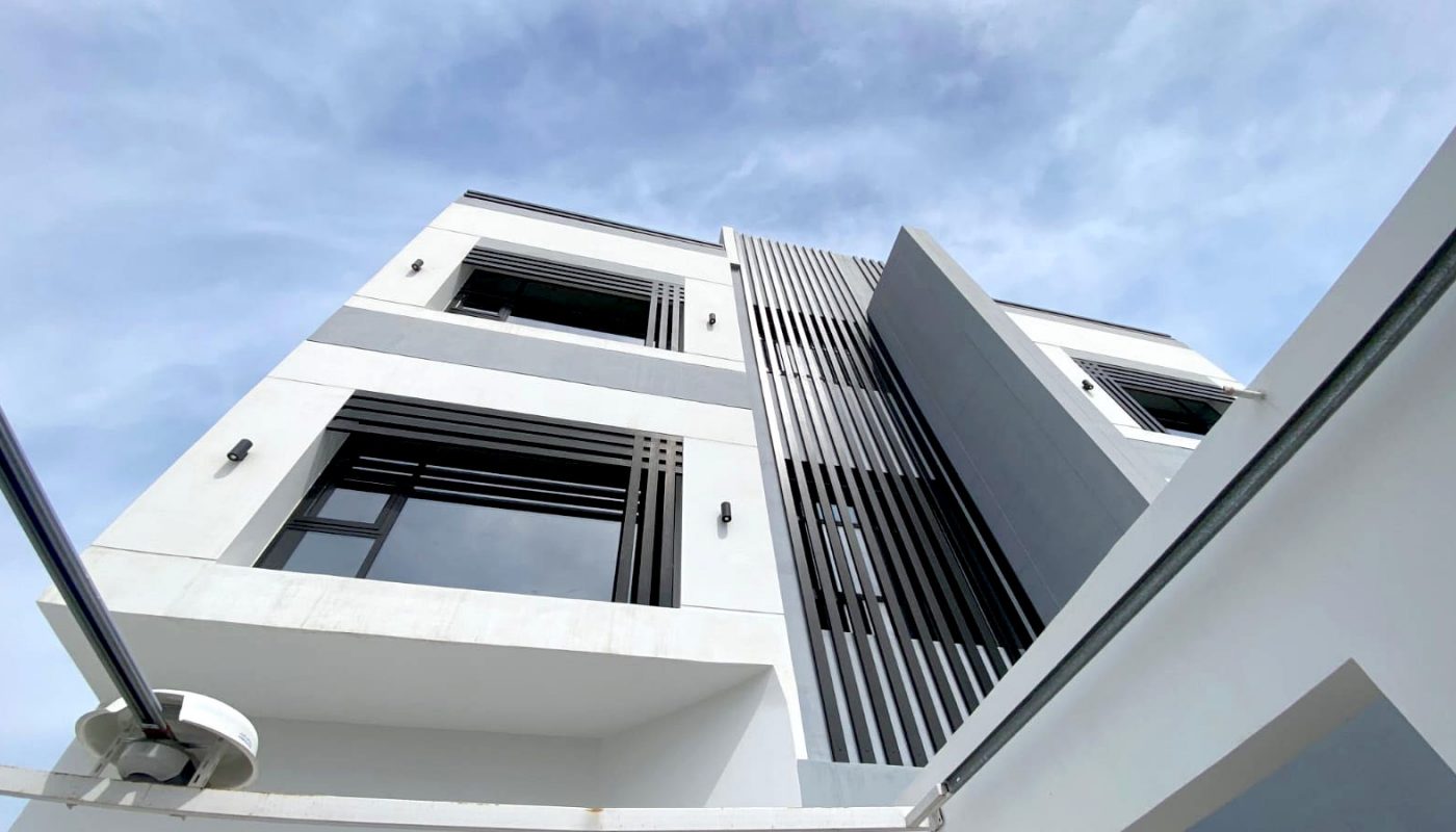 Low-angle view of a modern three-story building with large windows, vertical metal slats, and a white and gray exterior against a partly cloudy sky.