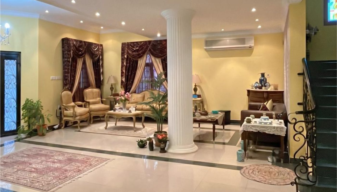 Spacious living room with beige armchairs, marble floors, a central white column, potted plants, a rug, and a staircase with ornate railing.