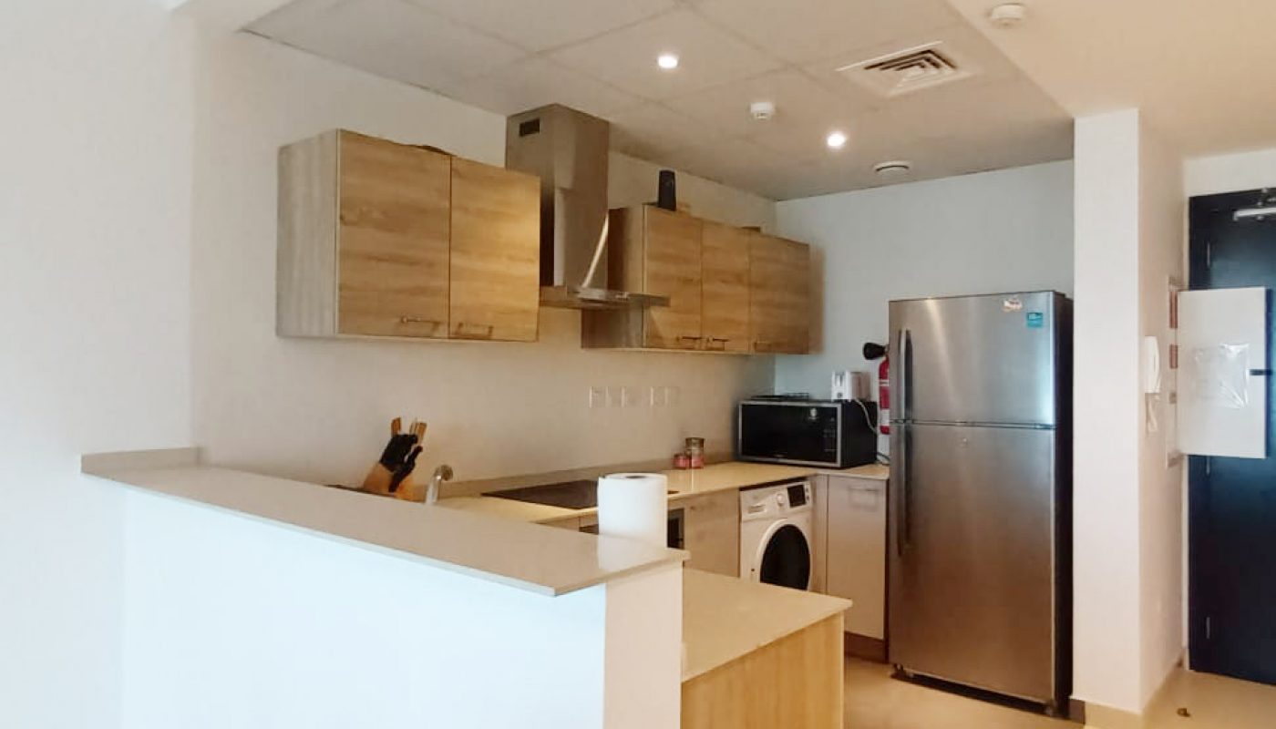 Modern kitchen with light wood cabinets, stainless steel appliances, a refrigerator, microwave, and trash bin. White countertops and walls provide a clean, minimal look.