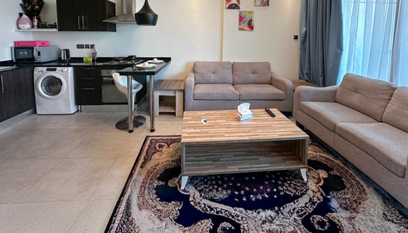 Modern open-plan living room with beige sofas, a wooden coffee table, patterned rug, kitchen area, dining table, chandelier, and large windows with sheer curtains.