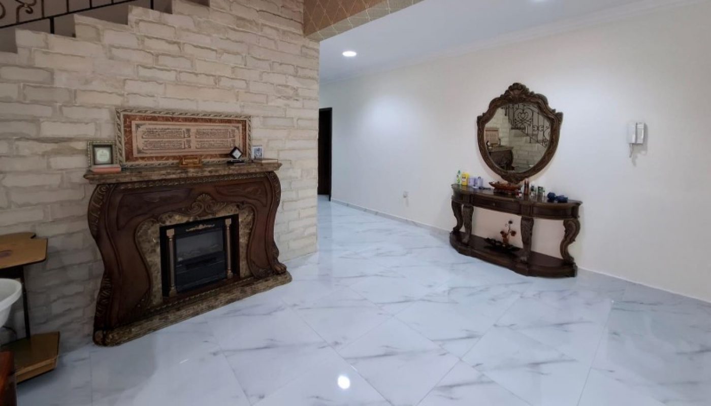Spacious room with white marble tile floor, ornate fireplace, stone wall, staircase with decorative railing, and a wooden console table with a mirror.