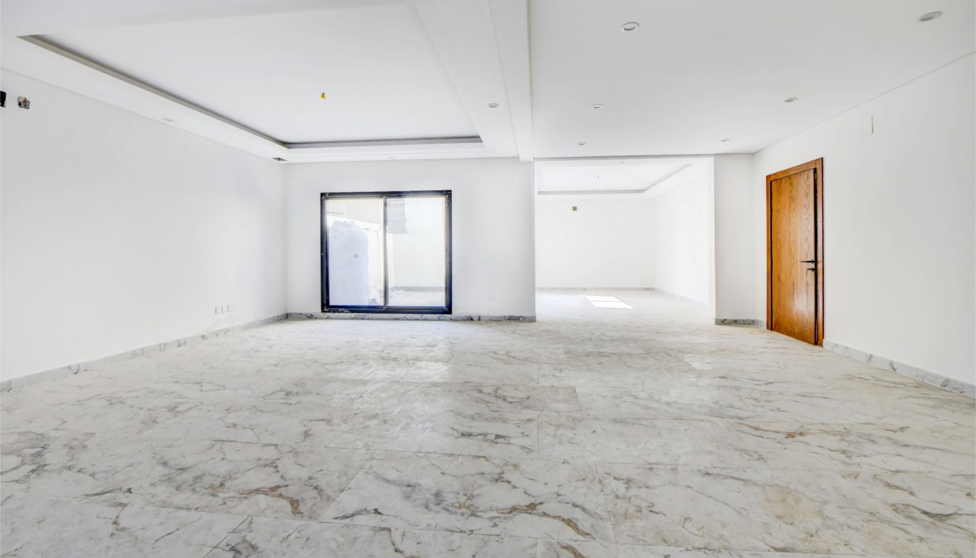 Empty room with white walls, marble floor, wooden door on the right, and large window at the back letting in natural light.