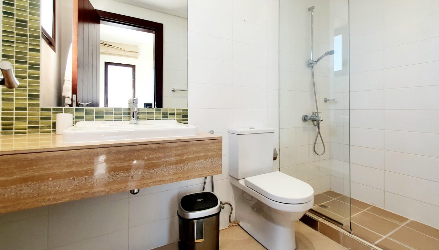 Modern bathroom with a sink, wall mirror, green tile backsplash, toilet, metal trash can, and a walk-in shower with glass enclosure and rain showerhead.