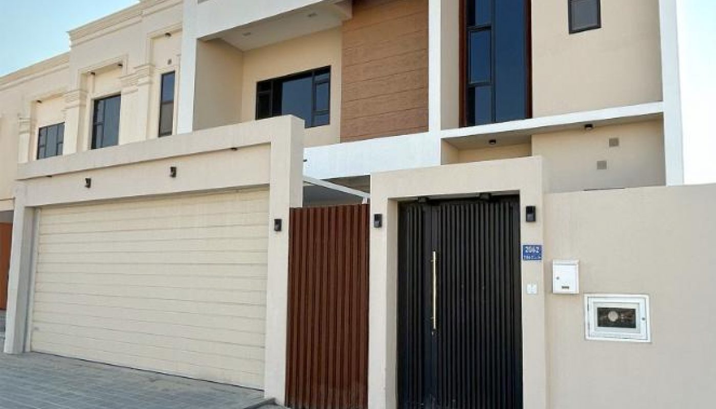 Modern two-story house with beige exterior, large garage door, black front gate, and rectangular architectural elements. Paved driveway in front.