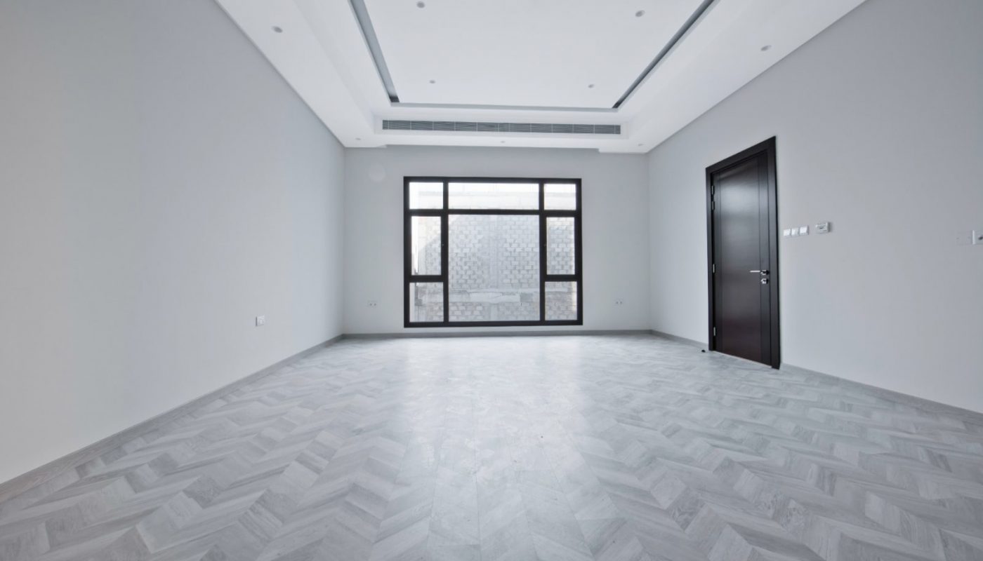 Empty room with herringbone parquet flooring, white walls, and large windows. A single dark door is on the right. Ceiling features recessed lighting and minimalist design elements.