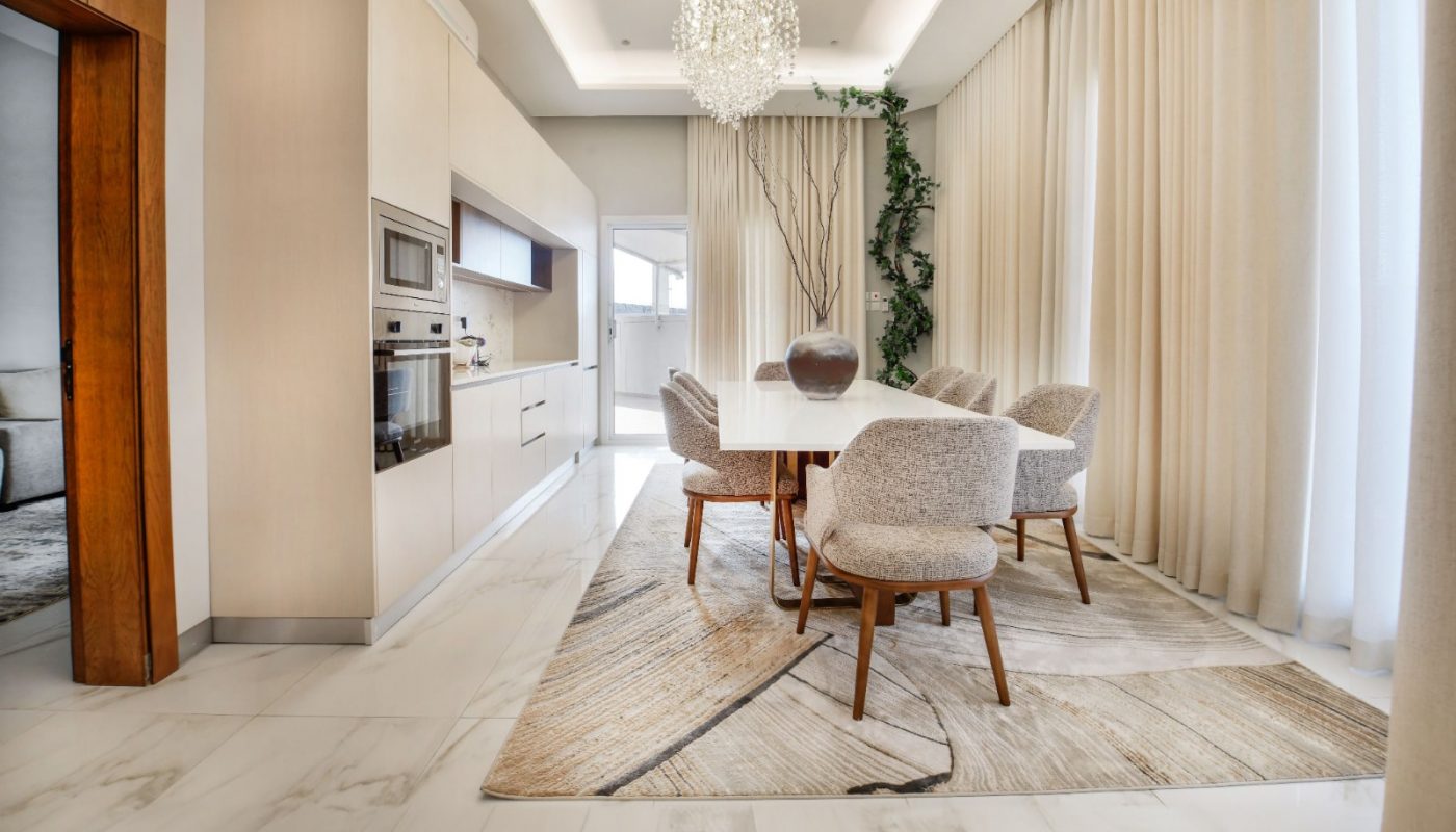 A modern dining room with a long table, six chairs, a chandelier, and large windows with curtains. The floor is tiled, and there's a plant in the corner.