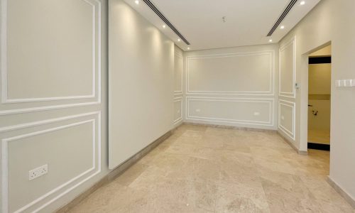 A spacious, empty room in an elegant villa for sale, featuring beige marble flooring, light gray walls with wainscoting, recessed lighting, and a doorway leading to another area. Located in the prestigious community of Diyar Al Muharraq.