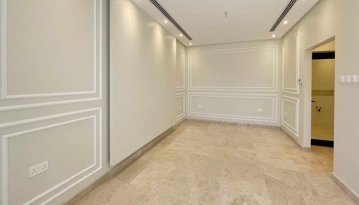 A spacious, empty room in an elegant villa for sale, featuring beige marble flooring, light gray walls with wainscoting, recessed lighting, and a doorway leading to another area. Located in the prestigious community of Diyar Al Muharraq.