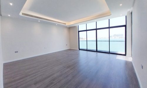 Spacious empty room with wooden flooring and large floor-to-ceiling windows revealing a water view. Ceiling features recessed lighting.