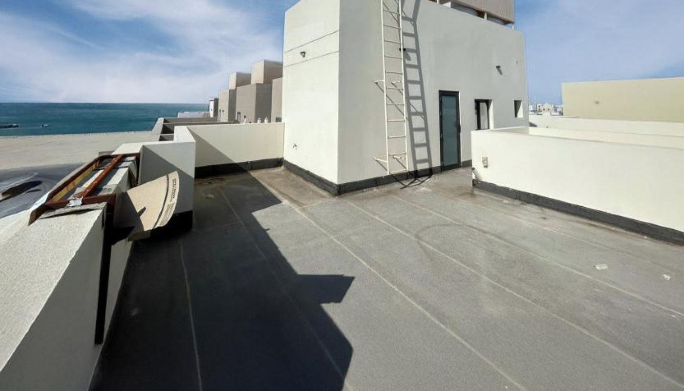 Flat rooftop with a view of the ocean, white modern building structures, a metal ladder, and blue sky with scattered clouds.