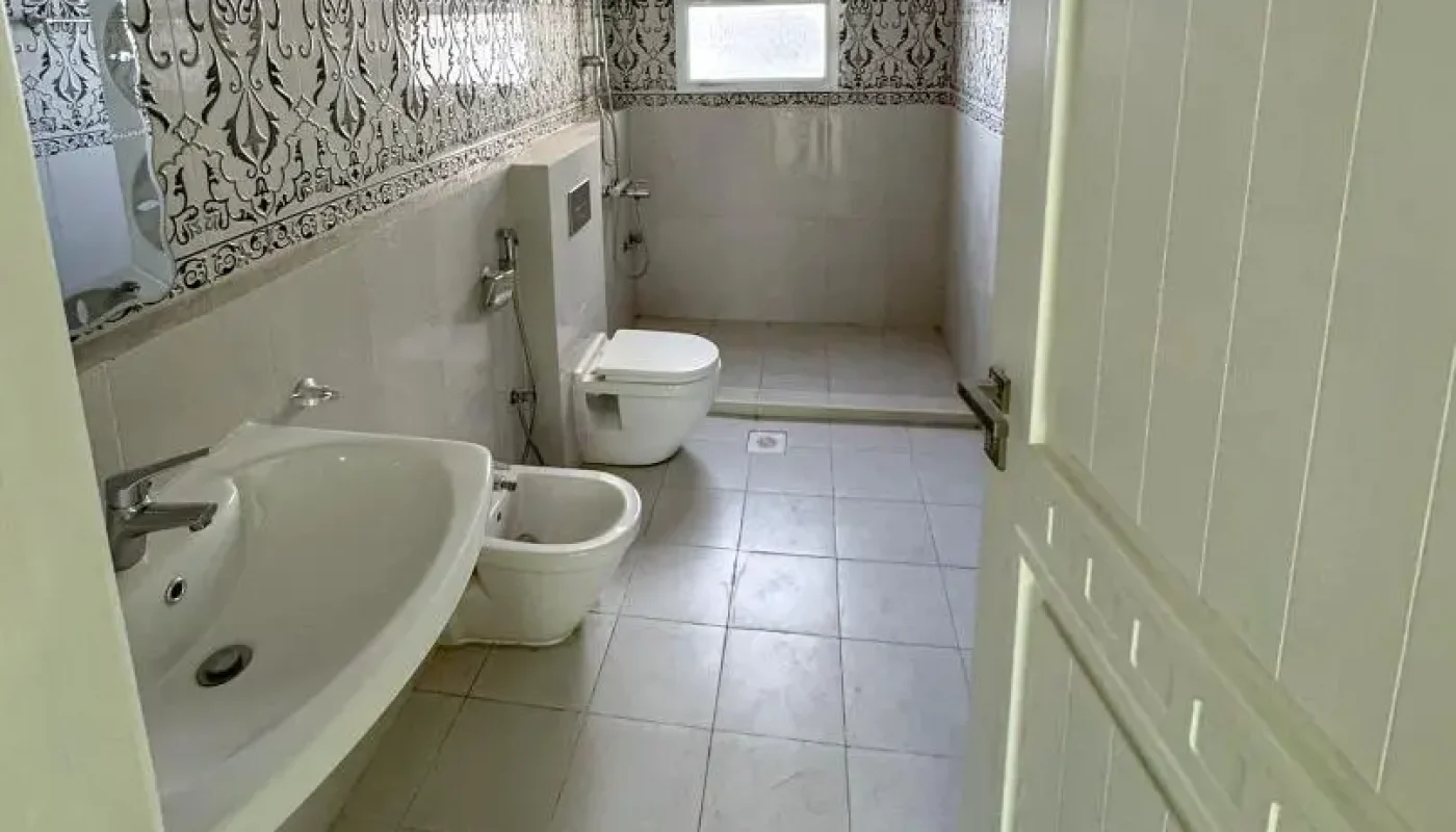 A bathroom with a sink, bidet, and toilet along one wall, decorative tile accents, a window, and a partially open white door.