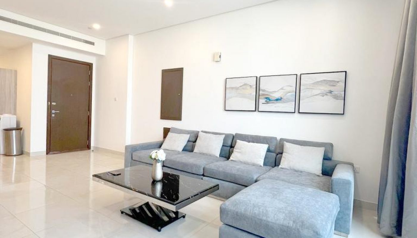 Modern living room with a gray sectional sofa, glass coffee table, white tile floor, minimal decor, and three framed artworks on the wall.