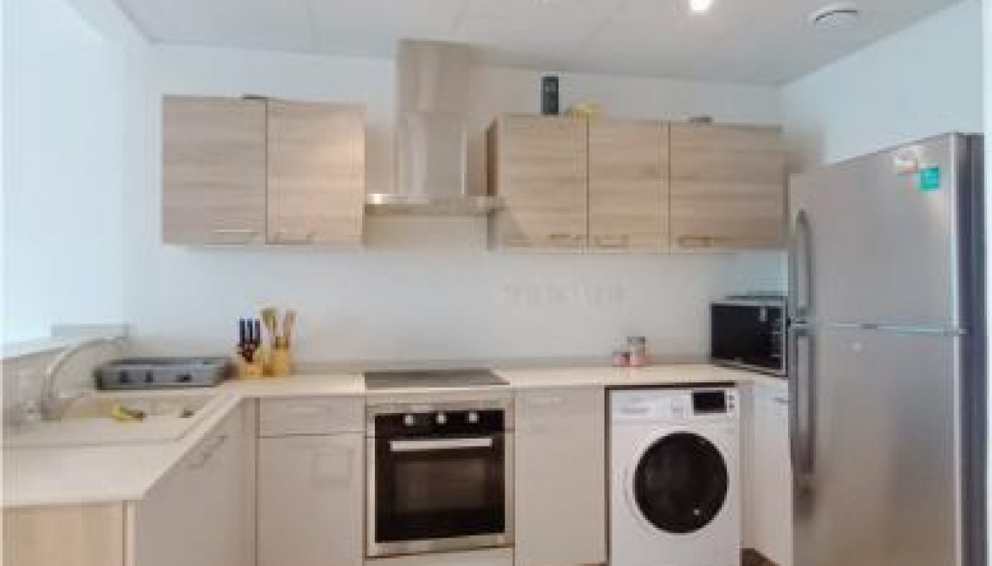 Modern kitchen with light wood cabinets, stainless steel oven, refrigerator, washing machine, and countertop appliances under bright ceiling lights.