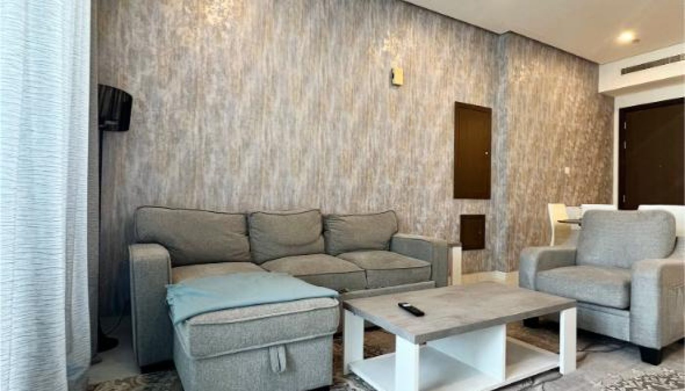 Modern living room with a gray sectional sofa, matching armchair, white coffee table, textured wallpaper, and patterned rug. A TV is mounted on the wall to the left.