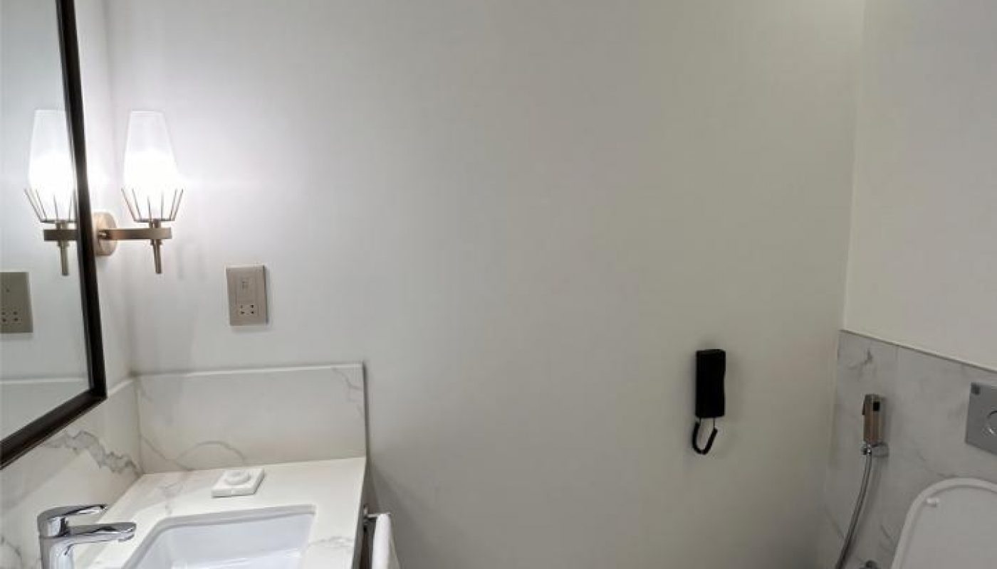 Modern bathroom with a white sink and countertop, wall-mounted light, tissue box, towel, toilet, bidet spray, phone, and waste bin on a white tiled floor and wall.