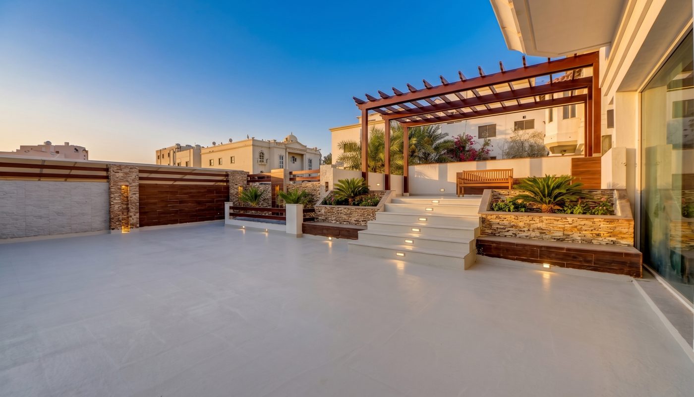 Modern patio with wide white tiled flooring, steps leading to a pergola, raised planters, palm plants, and residential buildings in the background under a clear blue sky.