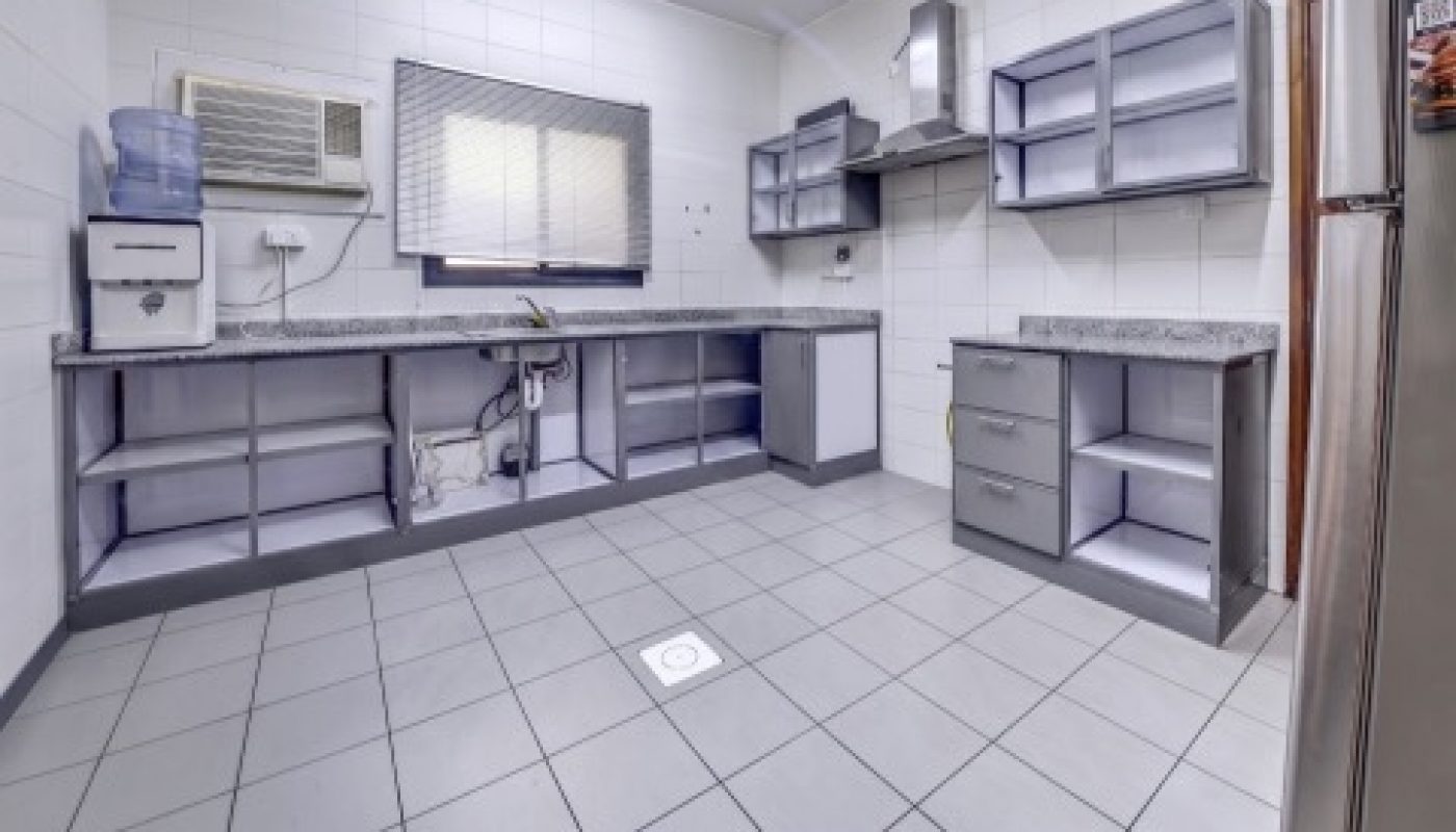 Empty kitchen with tiled floor, light gray cabinets, overhead fluorescent light, window with bars, water dispenser, and air conditioning unit on the left wall.