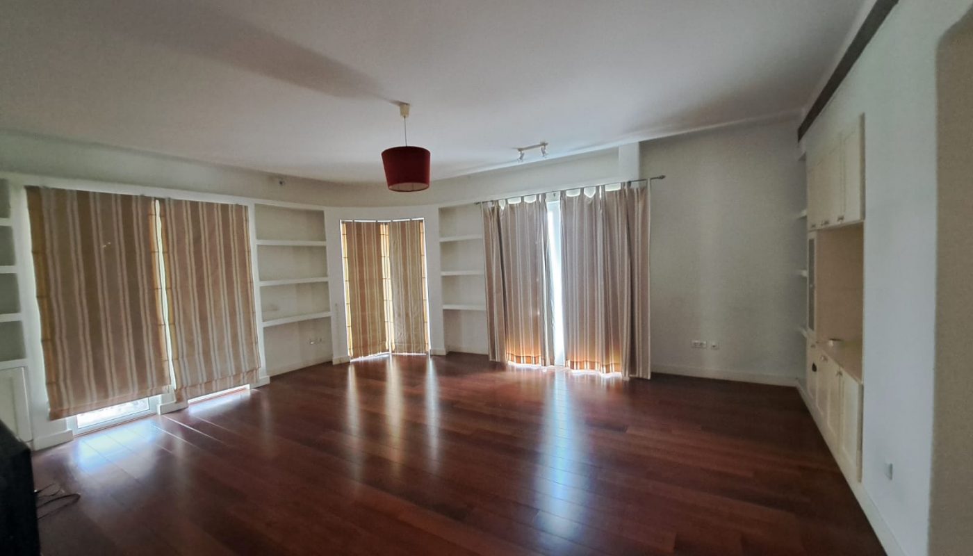 Spacious empty room with wooden flooring, large windows covered by beige curtains, built-in shelves on the walls, and a red ceiling light fixture.