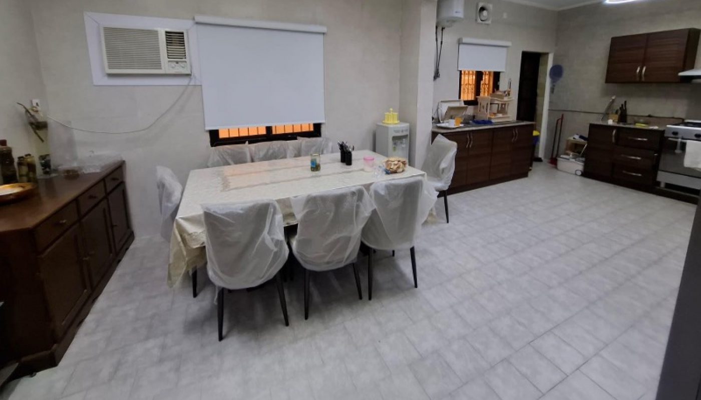 Spacious kitchen with a dining table, six covered chairs, wooden cabinets, tiled floor, modern light fixtures, and appliances along the walls.