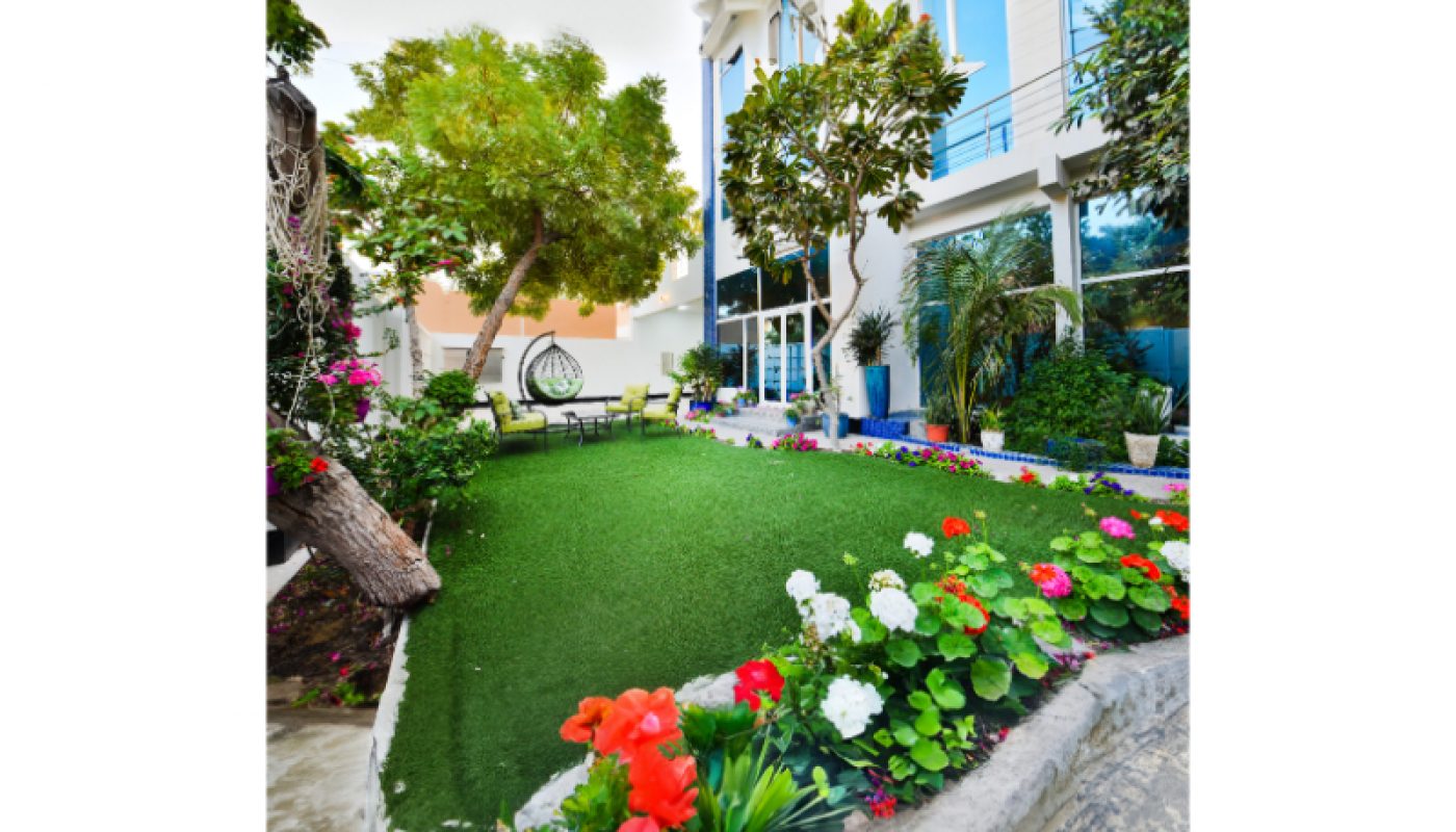 A landscaped garden with green grass, colorful flowers, trees, and outdoor seating next to a modern glass building.
