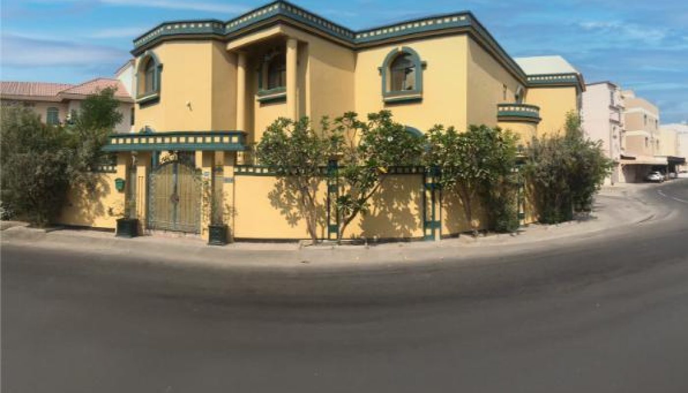 Two-story yellow house with green trim on a street corner, surrounded by a fence and trees, under a clear blue sky.