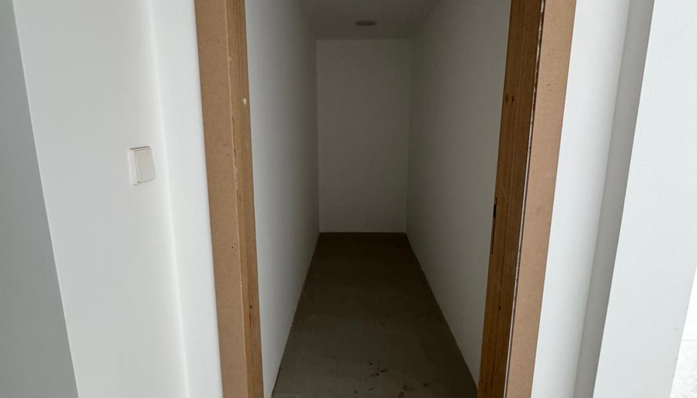 A narrow, empty room with white walls, a tiled floor, and a wooden door frame. A ceiling light and smoke detector are visible.