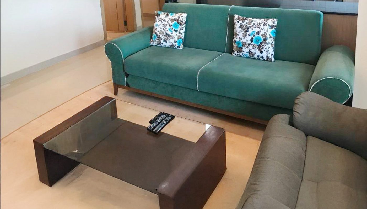 Modern living room with a green sofa, gray loveseat, glass coffee table, and open kitchen in the background. Two floral pillows are on the green sofa.