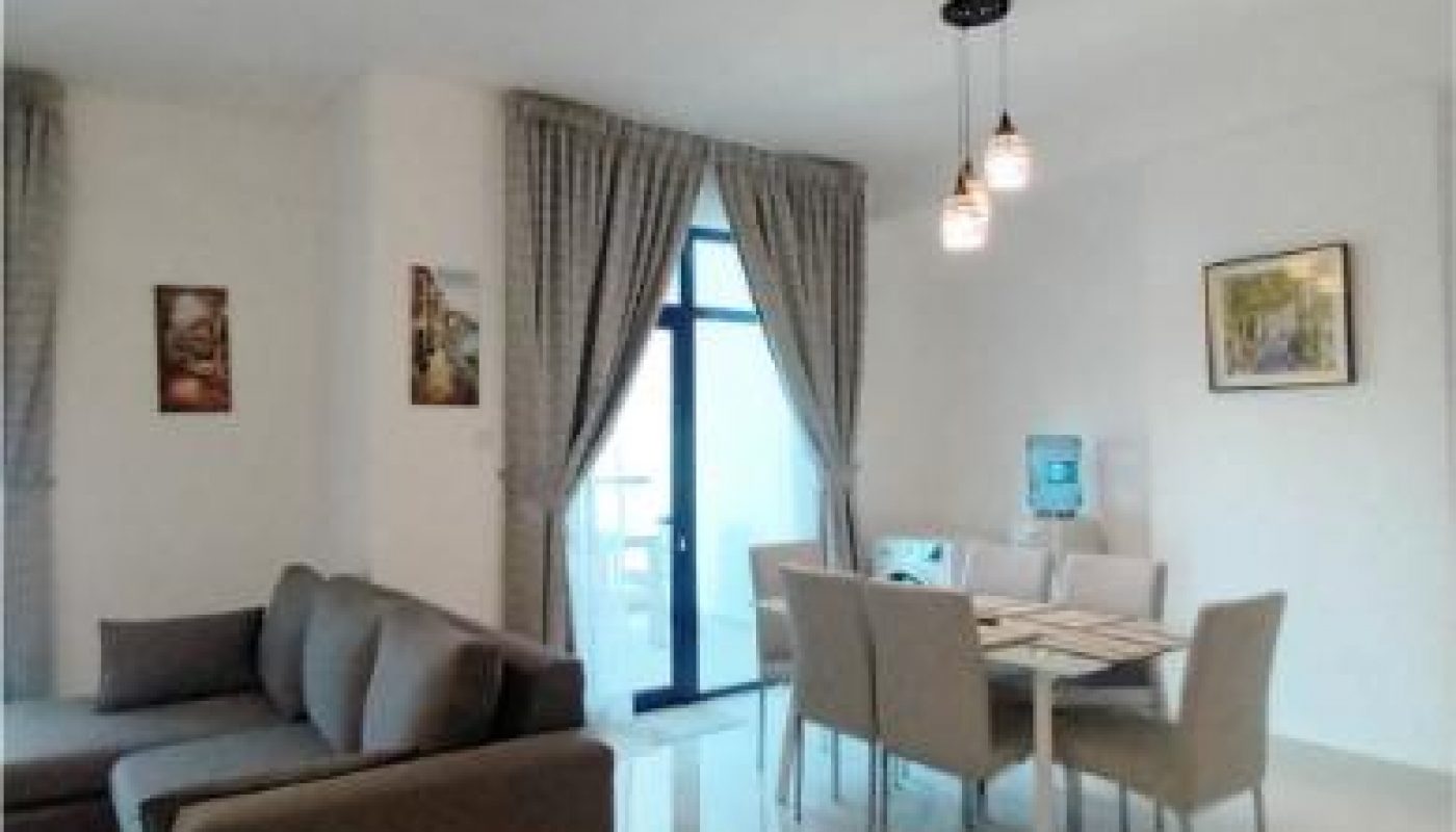 A modern living and dining area with a gray sofa, dining table with six chairs, pendant lights, and framed pictures on the walls. Large window with curtains lets in natural light.