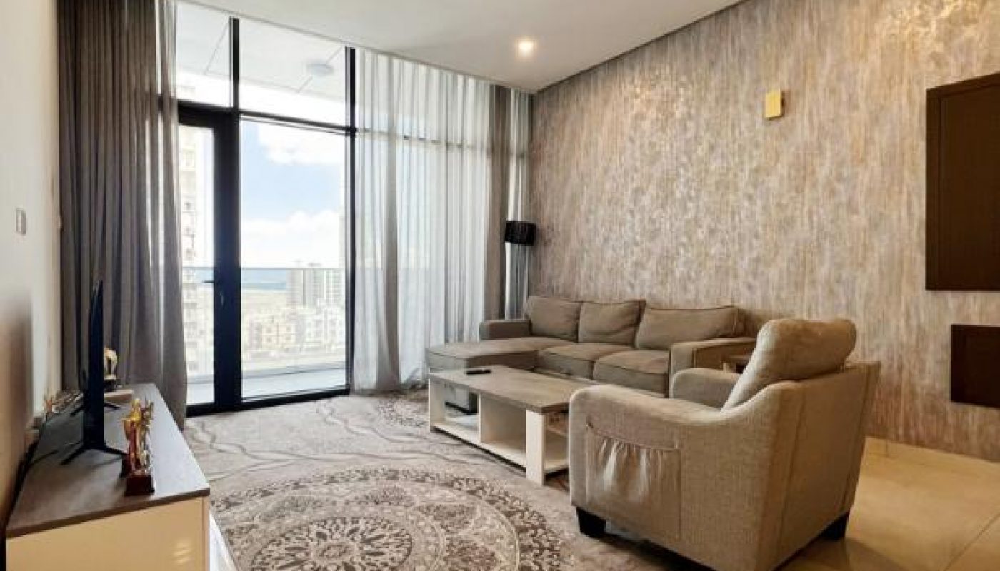 Modern living room with a gray sectional sofa, patterned rug, TV on a low stand, coffee table, large windows, and a textured accent wall.